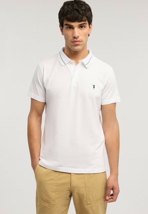 Giovane uomo con capelli ricci scuri, indossa una polo bianca a maniche corte e pantaloni beige, in piedi contro uno sfondo chiaro e uniforme.