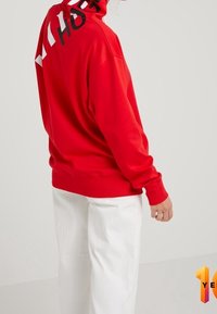 Persoon in een rode hoodie met zwarte en witte tekst op de capuchon, gecombineerd met losse witte broek, tegen een effen achtergrond.