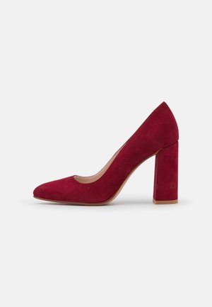 Zapato de tacón alto de ante rojo con puntera redondeada y tacón grueso. La parte superior presenta una textura lisa y un diseño de empeine bajo.