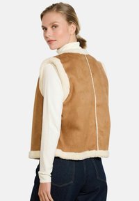 Sieviete ar blondiem matiem, tērpusies smilškrāsas zamša bezpiedurkņu vestē ar baltu šerpa apmalīti, zem kuras redzama balta turtlenecka un tumši zili džinsi, skatās atpakaļ.