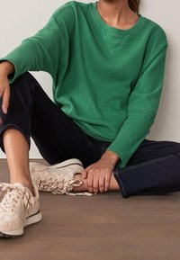 Chemise verte à manches longues texturée avec un col en V. Associée à un jean foncé et des baskets de couleur claire, dotées de lacets et d'une semelle rembourrée.