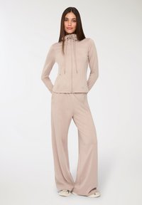 Giacca sportiva beige chiaro con cappuccio e zip frontale, abbinata a pantaloni beige larghi. Tessuto liscio e elasticizzato con dettagli minimi.