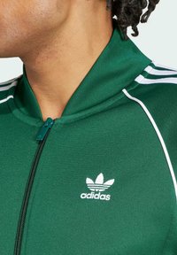 Chaqueta verde con cremallera y acentos blancos, con un alto cuello y diseño de tres rayas en los hombros. Incluye un logo blanco de Adidas en el pecho.