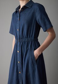 Denim skjortekjole med korte ermer, knap-lukning foran, elastisk talje, sidelommer og gullfargede knapper. Dyp blå farge og jevn tekstur.