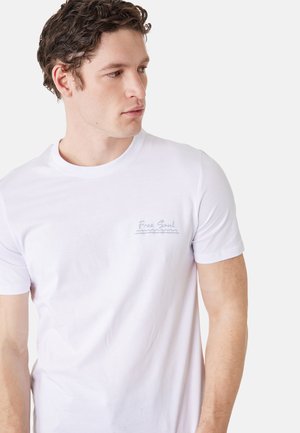 Hombre con cabello rizado que lleva una camiseta blanca con el texto "Free Soul" en el lado izquierdo del pecho, mirando hacia su derecha sobre un fondo liso.