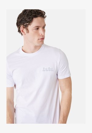 Mann mit lockigem Haar, der ein weißes T-Shirt mit dem Schriftzug „Free Soul“ auf der linken Brust trägt, schaut nach rechts vor einem einfarbigen Hintergrund.