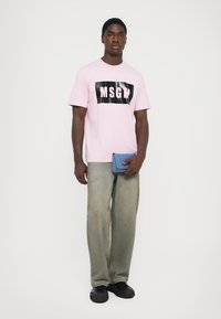 Lys rosa t-skjorte med svart "MSGM" logo, parret med falmede mørk grå jeans. Modellen holder en blå lommebok og bruker svarte sko.
