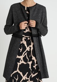 Žena upravuje texturovanou tmavě šedou bundu s dlouhým rukávem přes černé a béžové šaty s abstraktním vzorem.