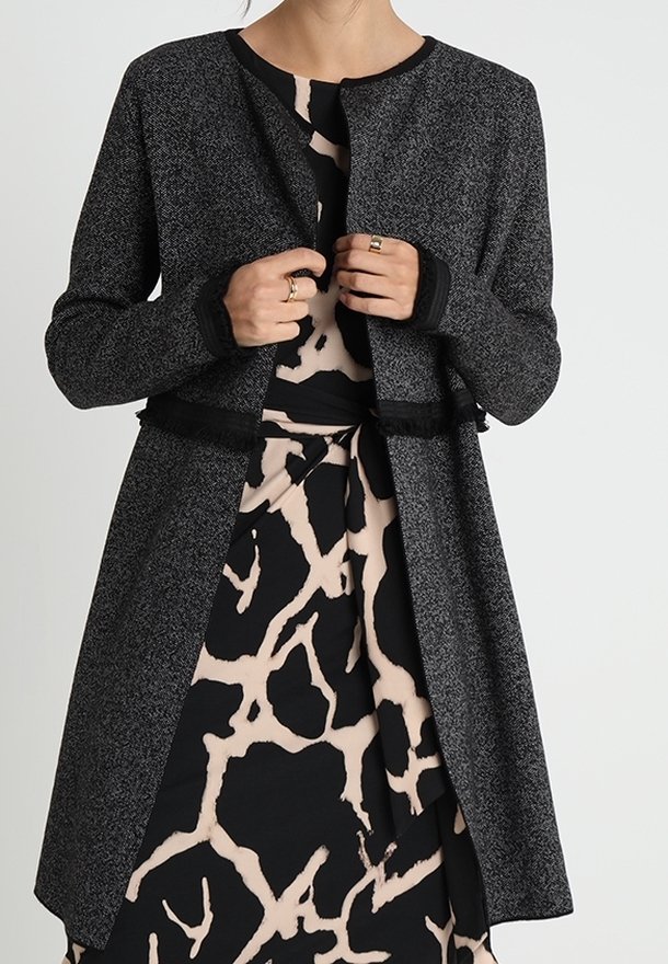 Žena upravuje texturovanou tmavě šedou bundu s dlouhým rukávem přes černé a béžové šaty s abstraktním vzorem.