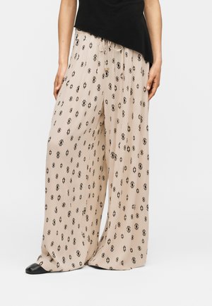 Beige bukser med vide ben og sort geometrisk mønster, snørretalje, båret med en sort top og sorte sko på en hvid baggrund.