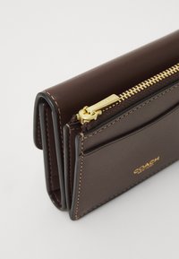 Braune Lederbörse mit goldfarbener Reißverschlussdetail, ausgestattet mit Kartenfächern und dezenten Nähakzenten. Kompakte Bauweise, glatte Textur.