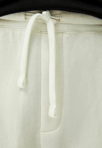 Detailní záběr na zavázanou bílou šňůrku na světle béžových teplácích s viditelným stehem a černým pasem.