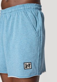 Gaiši zili kokvilnas šorti ar elastīgu jostu, sānu kabatām un melnu Nike logo uzšuvumu apakšējā kreisajā stūrī. Iezīme ir neapstrādātā apmale.