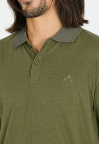 Olivgrön pikétröja i mjukt material med en kontrasterande mörkgrön krage, med en liten logotyp i en ljusare nyans. Tvåknappsstängning.