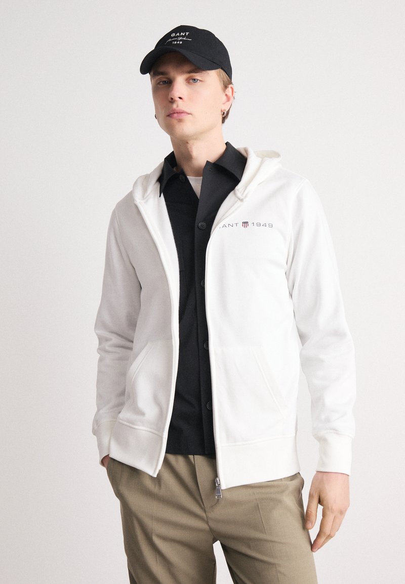 Felpa bianca con zip e cappuccio, tasche frontali e logo "GANT 1949". Abbinata a una camicia nera con bottoni e pantaloni beige.
