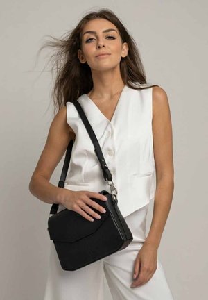 Černá kožená crossbody taška s klopou, měkkou texturou, geometrickým designem, stříbrnými detaily a odnímatelným popruhem, držena jednou rukou.