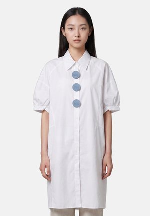 Femme portant une robe-chemise blanche mi-longue avec des manches courtes bouffantes et trois gros boutons ronds bleus sur le devant.