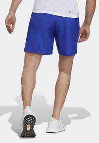 Man med blå sportshorts, vita sneakers och vit t-shirt, som går bort med synlig skugga på en ljus bakgrund.