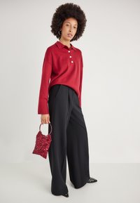 Polo de malha vermelho com três botões, combinado com calças largas pretas. A segurar uma pequena mala burdeos com lantejoulas e uma alça circular.