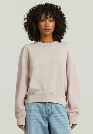 Woman with curly black hair wearing a light beige long-sleeve sweatshirt and light blue jeans, standing against a plain background.