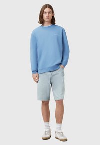 Světle modrý svetr s kulatým výstřihem a rozšířenými rukávy, v kombinaci se světle modrými džíny. Model má na sobě bílé ponožky a ležérní tenisky.