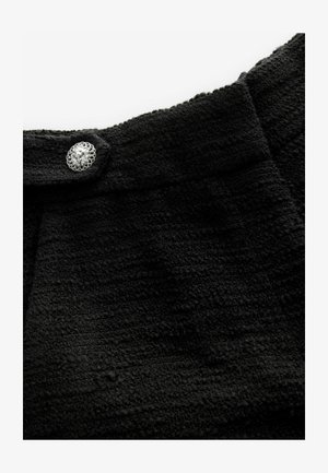 Tisă neagră cu textură, cu un model țesut și un buton decorativ argintiu cu un design floral aproape de decolteu.