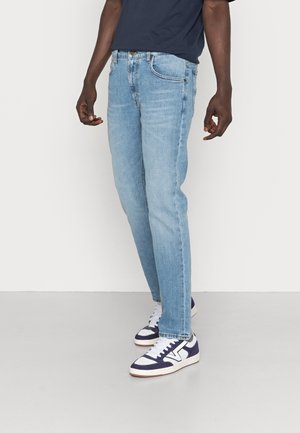 Jeans de denim azul claro con un corte recto, que cuentan con bolsillos frontales y un cierre de botón clásico, combinados con zapatillas de deporte en navy y blanco.