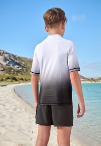 Kortärmad rashguard med en gradient från vitt till svart, med ett prickmönster. Matchas med svarta badshorts. Strandmiljö.
