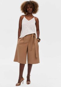 Witte, mouwloze, gebreide top met een v-hals en uitgesneden patroon, gecombineerd met hoog-getailleerde, tan kleurige culotte broeken die strakker zijn bij de taille. Bruine hakken.