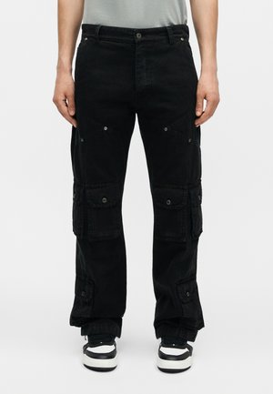 Homme portant un pantalon cargo noir avec plusieurs poches à rabat et des baskets noir et blanc, debout devant un fond uni.