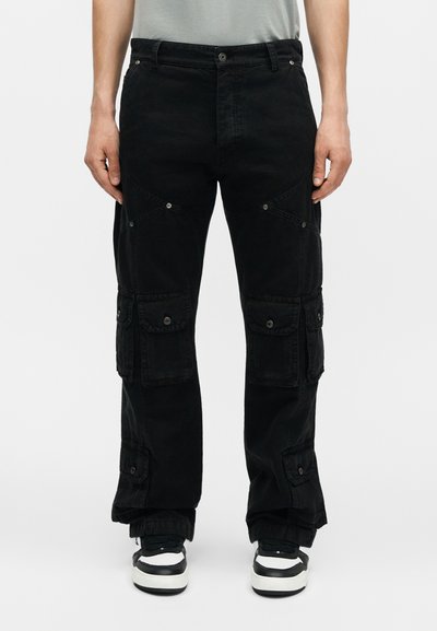 Homme portant un pantalon cargo noir avec plusieurs poches à rabat et des baskets noir et blanc, debout devant un fond uni.