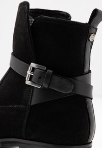 Stivale alla caviglia in suede nero con cinturino in pelle e dettaglio in metallo argentato, caratterizzato da cuciture testurizzate e una linguetta sul retro.
