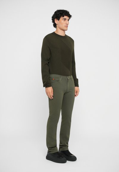 Olíva zöld pulóver, texturált, átlós mintázattal, illesztve hozzá illő olíva zöld, slim-fit farmernadrággal és fekete sportcipőkkel.