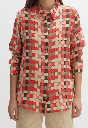 Femme portant une chemise boutonnée à manches longues avec un motif géométrique de rectangles rouges, beiges, marron et noirs sur un pantalon en velours côtelé beige.