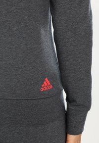 Ciemnoszara bluza wykonana z miękkiego materiału, z widocznym czerwonym logo Adidas na dolnym brzegu. Projekt obejmuje dopasowany krój.