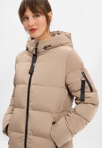 Beige Steppjacke mit Kapuze, gesteppter Optik, schwarzem Reißverschluss und Seitentaschen. Verfügt über einen schwarzen Akzent am Reißverschluss und ein Schlaufen-Detail.