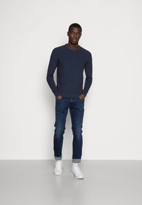 Pull en maille bleu marine avec un col rond, associé à un jean bleu clair et des baskets blanches. Le tissu semble lisse avec des détails de poignets côtelés.