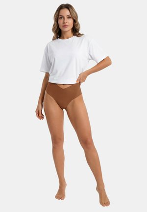 Vrouw staat op blote voeten, draagt een wit topje met korte mouwen en hoogtalige bruine onderkleding, met de hand rustend op de taille.