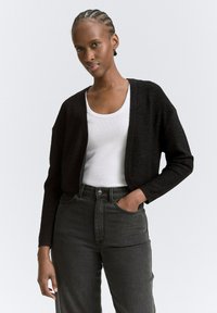 Schwarze Strickjacke mit lockerer Passform und offenem Front, getragen mit einem weißen Tanktop und dunklen, hochgeschnittenen Jeans.