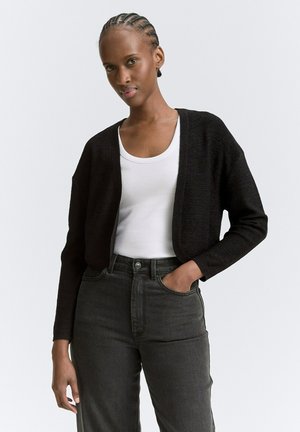 Zwarte gebreide cardigan met een losse pasvorm en open voorkant, gecombineerd met een witte tanktop en donkere hoogwaisted jeans.