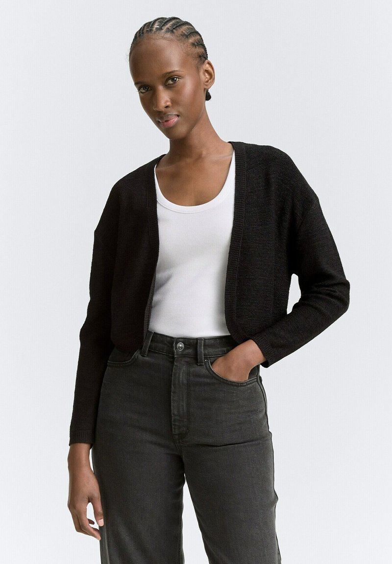 Schwarze Strickjacke mit lockerer Passform und offenem Front, getragen mit einem weißen Tanktop und dunklen, hochgeschnittenen Jeans.