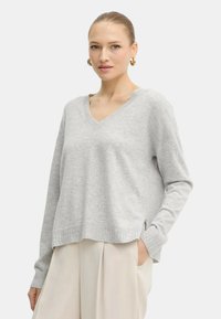 Donna con capelli raccolti che indossa orecchini a cerchio dorati, un maglione leggero grigio V-neck e pantaloni larghi color crema con una mano in tasca.