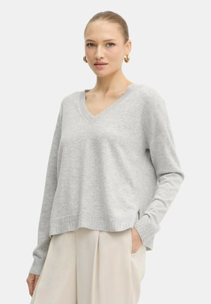 Donna con capelli raccolti che indossa orecchini a cerchio dorati, un maglione leggero grigio V-neck e pantaloni larghi color crema con una mano in tasca.