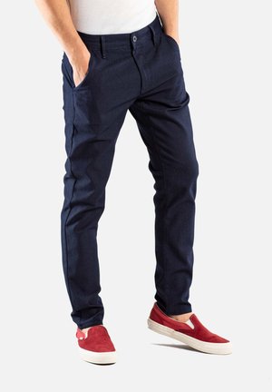 Marineblaue Stretchhosen mit schmaler Passform, ausgestattet mit Seitentaschen und einem Knopfverschluss. Komplementiert mit roten Slippern.