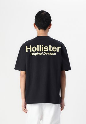 Mand vender væk iført en sort t-shirt med stor gul tekst "Hollister Original Designs" på ryggen og hvide bukser.