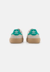 Beige suede sneakers with a textured gum sole and green accent heel tab featuring white Adidas logo. Classic low-top design.
