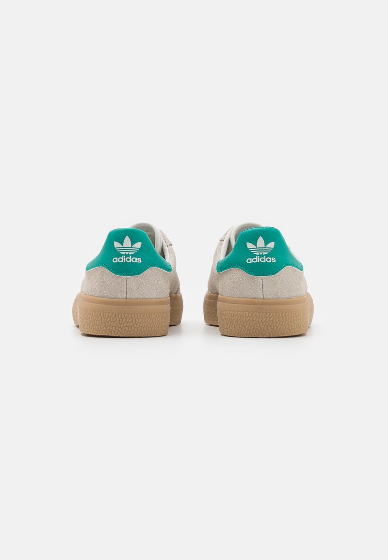 Baskets en daim beige avec une semelle en gomme texturée et un onglet de talon vert arborant le logo Adidas blanc. Design classique à tige basse.