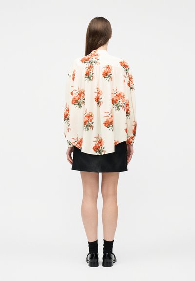 Γυναίκα με την πλάτη γυρισμένη, φοράει λευκό πουκάμισο με πορτοκαλί λουλουδένιο σχέδιο, μαύρη φούστα, μαύρες κάλτσες και μαύρα παπούτσια, μπροστά σε απλό φόντο.