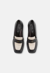 Černé kožené mokasíny s krémovým koženým panelem, čtveratou špičkou a dekorativním páskem s kovovým detailem. Viditelná texturovaná podešev a podšívka.