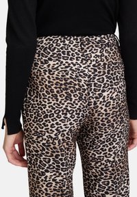 Los pantalones de estampado de leopardo tienen una base beige clara con manchas negro-marrón oscuro. Incluyen bolsillos traseros y una cinturilla ajustada. Parte superior negra visible.